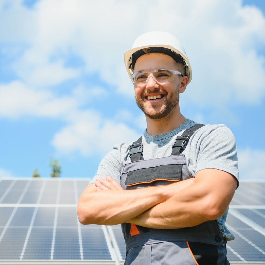 Solar panel technician in a hard hat and safety goggles standing in front of photovoltaic panels with arms crossed, smiling.