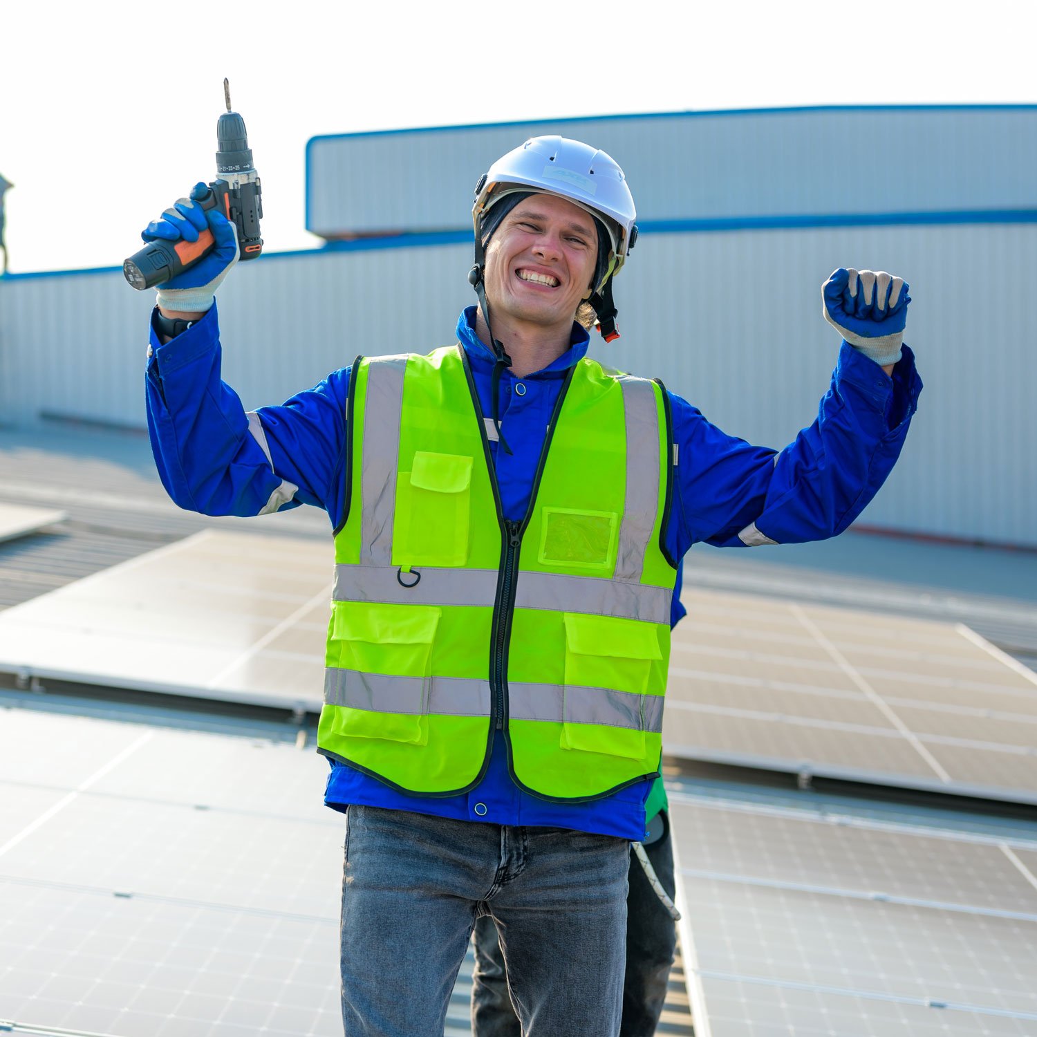 Worker on a rooftop in a neon safety vest and hard hat, holding a cordless drill and smiling beside solar panels.